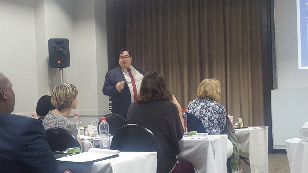 Howard Fox A man in a suit and tie addresses a group of people sitting at desks.