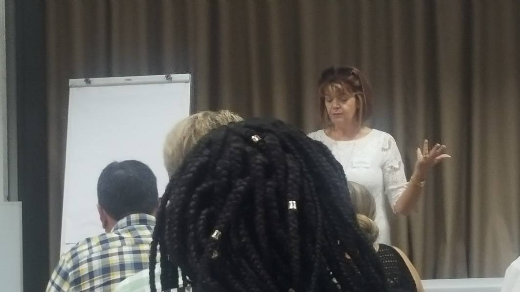Marina Short A woman in a white shirt addresses three people, seated in rows.