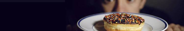 Temptation to tamper A close up of a donut with chocolate icing and rainbow sprinkles on a plate, with a man's wide-eyed face in the background.
