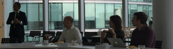 Meet Max Kantelia.fw Three people sit and one man stands at a boardroom desk.