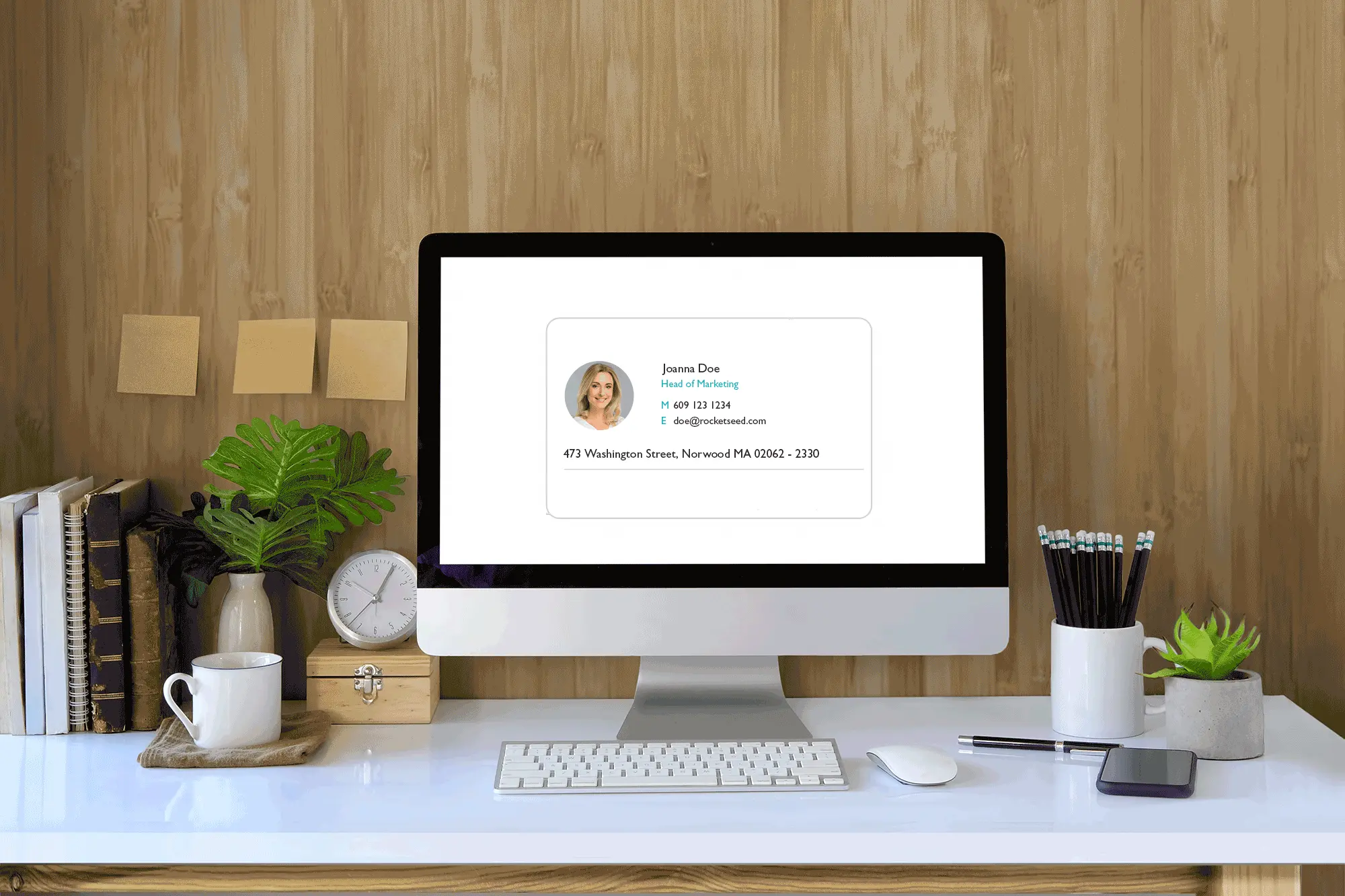 Your new email signature manager A desktop showing a computer screen with an email signature for "Joanna Doe, Head of Marketing," including her contact details. The desk has books, a plant, a clock, and pencils.