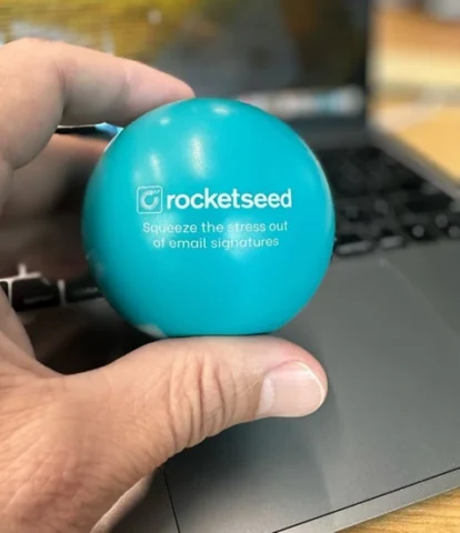 Rocketseed Stress ball A hand holding a blue Rocketseed stress ball that says "squeeze the stress out of email signature management" with a laptop background